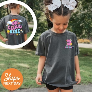 May include: A dark gray t-shirt with the text "ON CLOUD NINE" and a rainbow, cloud, and smiley face design on the back. The front of the shirt has a pink number 9 with a smiley face and the name "AUDREY".