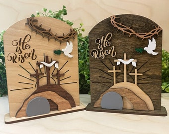 Puede incluir: Dos decoraciones de Pascua de madera con las palabras "He is Risen" en cada una. Las decoraciones presentan una cruz, una paloma y una tumba de piedra.