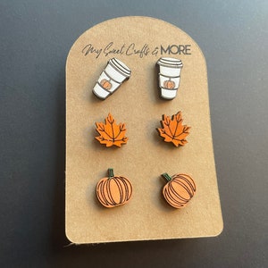 May include: A set of six wooden earrings featuring fall-themed designs. The earrings include two pumpkin designs, two maple leaf designs, and two coffee cup designs with pumpkins inside. The earrings are displayed on a brown card with the text "My Sweet Crafts & MORE".