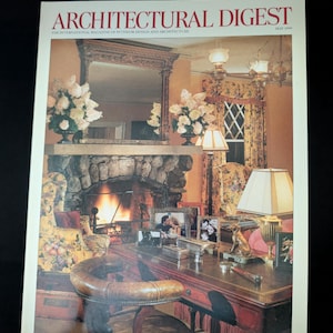 Może przedstawiać: Okładka magazynu Architectural Digest, maj 1999, przedstawia luksusowy wystrój wnętrz. Widoczny jest kominek z płonącym ogniem, biurko z oprawionymi zdjęciami i skórzany fotel. Tytuł magazynu jest czerwony.
