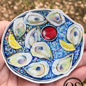 May include: A sticker of a plate of oysters on a bed of blue glitter. The oysters are arranged in a circle around a small dish of red sauce. There are two lemon wedges on the plate. The sticker has the text "Queenstown Studios" on the bottom right corner.