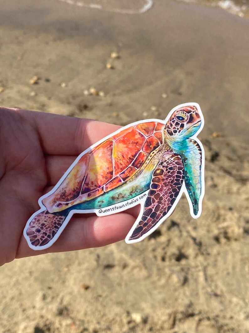 May include: A colorful sticker of a sea turtle with a vibrant orange, yellow, and blue shell. The sticker has a white border and the text "Queenstownstudios.com" is printed on the bottom.