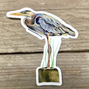 May include: A watercolor sticker of a blue heron standing on a wooden post with green reeds in the background. The sticker has the text "Queenstownstudios.com" on the bottom left corner.