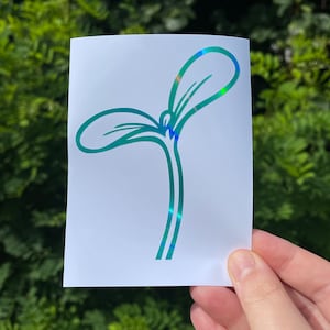 May include: A white sticker featuring a teal-colored sprout design with iridescent accents. The sprout has two leaves and a stem, and the sticker is held against a backdrop of green foliage.