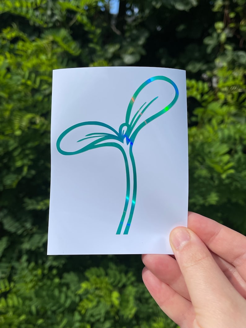 May include: A white sticker featuring a teal-colored sprout design with iridescent accents. The sprout has two leaves and a stem, and the sticker is held against a backdrop of green foliage.