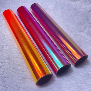 May include: Three rolls of iridescent vinyl film in shades of orange, pink, and purple. The rolls are lying on a white fuzzy surface.