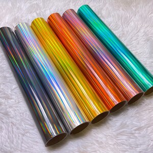 May include: A collection of seven rolls of holographic vinyl in various colors, including silver, gold, orange, pink, and teal. The vinyl has a reflective, iridescent finish, creating a shimmering effect. The rolls are arranged diagonally, showcasing the range of colors.