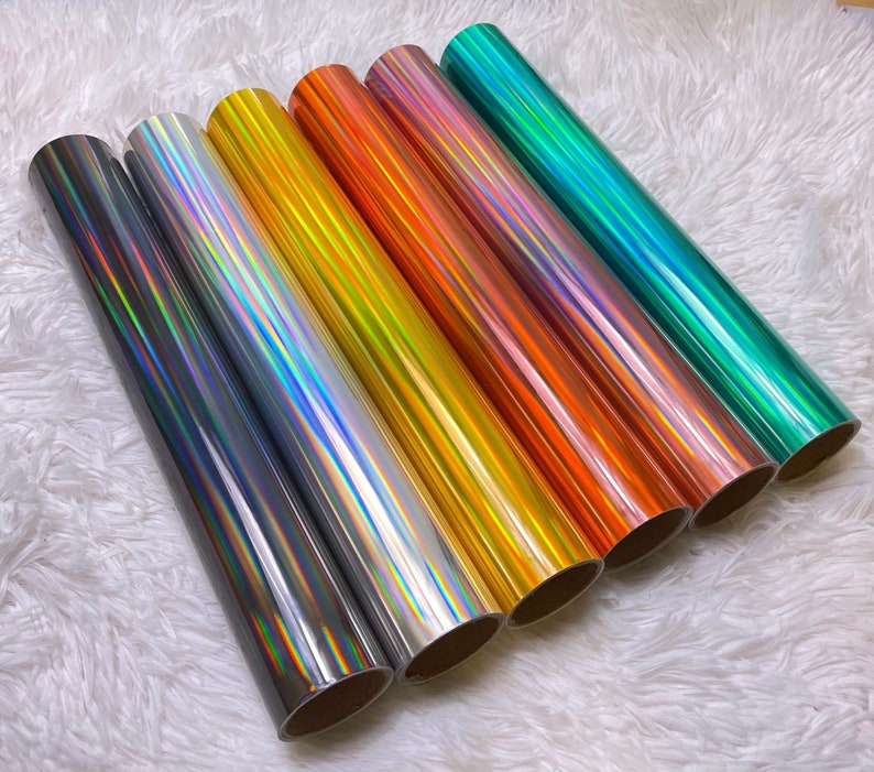 May include: A collection of seven rolls of holographic vinyl in various colors, including silver, gold, orange, pink, and teal. The vinyl has a reflective, iridescent finish, creating a shimmering effect. The rolls are arranged diagonally, showcasing the range of colors.
