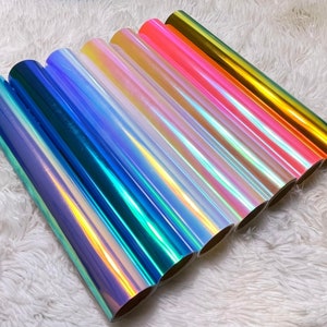 May include: A collection of iridescent vinyl rolls in various colors, including blue, green, pink, and gold. The rolls are arranged diagonally, showcasing their shiny, color-shifting surfaces. The background is a soft, white, textured fabric.