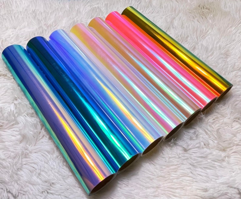 May include: A collection of iridescent vinyl rolls in various colors, including blue, green, pink, and gold. The rolls are arranged diagonally, showcasing their shiny, color-shifting surfaces. The background is a soft, white, textured fabric.