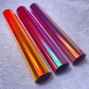 May include: Three rolls of iridescent vinyl in shades of orange, red, and purple. The vinyl has a glossy finish and reflects multiple colors. The rolls are arranged side by side on a white, fluffy surface.