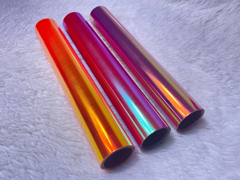 May include: Three rolls of iridescent vinyl in shades of orange, red, and purple. The vinyl has a glossy finish and reflects multiple colors. The rolls are arranged side by side on a white, fluffy surface.