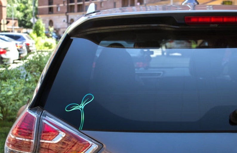 May include: A teal and blue sprout sticker is affixed to the rear window of a car. The sticker features a minimalist design of a young plant with two leaves and a stem, suggesting growth and nature.