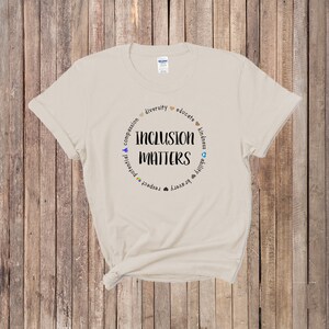 May include: A beige t-shirt with a circle graphic that says "Inclusion Matters" surrounded by words like "diversity", "educate", "compassion", "potential", "respect", "bravery", "ability", "kindness", and "unity".