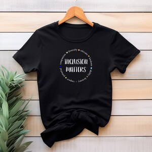 May include: Black t-shirt with a white circle graphic that says "Inclusion Matters" surrounded by the words "compassion, diversity, educate, kindness, ability, respect, potential, equity, unity".