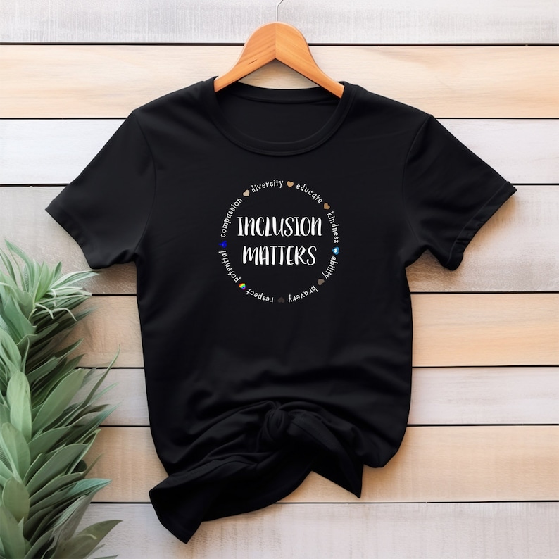 May include: Black t-shirt with a white circle graphic that says "Inclusion Matters" surrounded by the words "compassion, diversity, educate, kindness, ability, respect, potential, equity, unity".