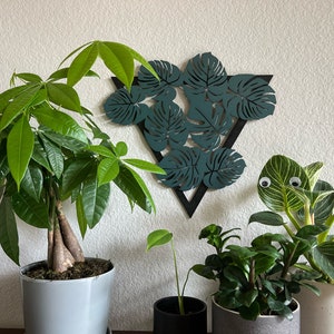May include: Three potted plants, a money tree, a monstera, and a prayer plant, with a teal green geometric wall decor featuring a triangle shape with cut-out monstera leaves.