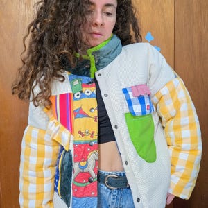 May include: A quilted jacket with a patchwork design. The jacket features a cream-colored body with a small checkered pattern, yellow and white checkered sleeves, and colorful patches with toy-themed illustrations. It has a green collar and a black button closure.