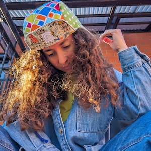 May include: A person wearing a colourful, patterned hat with a city scene design. The hat has a green, orange, blue and white geometric pattern. The person is wearing a blue denim shirt and jeans.
