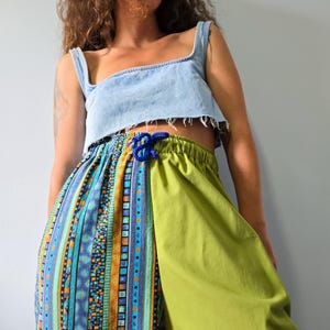 May include: A light blue denim crop top with a raw hem paired with loose-fitting trousers. The trousers feature a split design with one side in solid lime green and the other side in a multi-coloured vertical striped pattern with blue, orange, and teal.