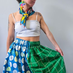 May include: A light gray spaghetti strap crop top paired with a two-toned skirt featuring a blue side with white and lime green circular patterns and a green side with a white grid pattern, accented by a colorful patterned neck scarf and earrings.