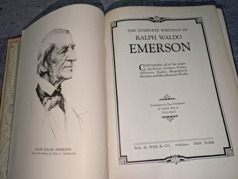 May include: Open book with a portrait of Ralph Waldo Emerson and the title "The Complete Writings of Ralph Waldo Emerson." The book is Volume 1, published by WM. H. Wise & Co. in New York. The book's pages are off-white.