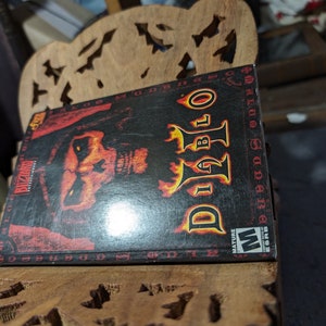 Op de afbeelding: Een zwarte videospeldoos met een rood en geel Diablo-logo en een demonisch gezicht op de voorkant. De doos is gelabeld "Blizzard Entertainment" en heeft een "Mature"-classificatie van de ESRB.