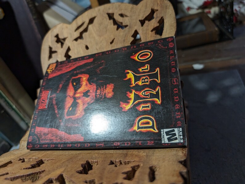 Op de afbeelding: Een zwarte videospeldoos met een rood en geel Diablo-logo en een demonisch gezicht op de voorkant. De doos is gelabeld "Blizzard Entertainment" en heeft een "Mature"-classificatie van de ESRB.