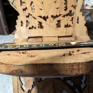 Op de afbeelding: Een houten stoel met ingewikkelde houtsnijwerk, met een boek dat op de zitting ligt. De stoel is gemaakt van lichtbruin hout en heeft een uniek ontwerp.