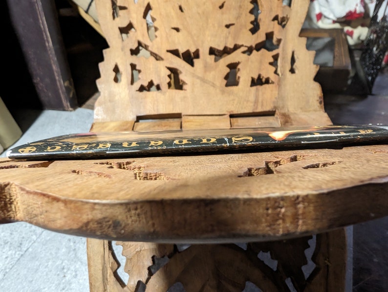 Op de afbeelding: Een houten stoel met ingewikkelde houtsnijwerk, met een boek dat op de zitting ligt. De stoel is gemaakt van lichtbruin hout en heeft een uniek ontwerp.