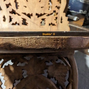Op de afbeelding: Een close-up van een houten tafel met ingewikkelde houtsnijwerk. De tafel heeft een glazen blad met de tekst "Diablo" II erop gedrukt.