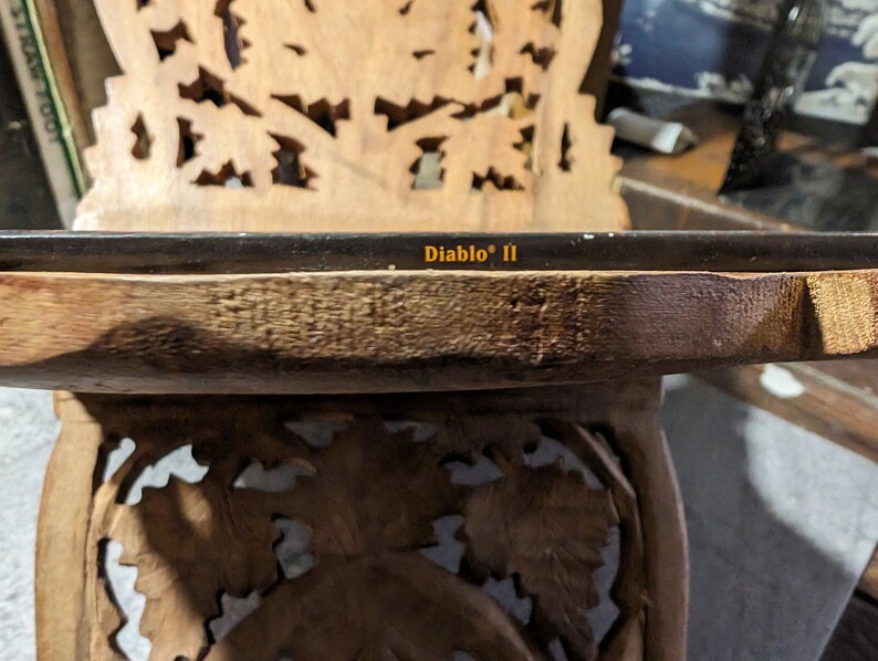 Op de afbeelding: Een close-up van een houten tafel met ingewikkelde houtsnijwerk. De tafel heeft een glazen blad met de tekst "Diablo" II erop gedrukt.
