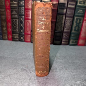 May include: A vintage, brown leather-bound book titled "The Works of Maupassant" with gold lettering. The book has a worn, textured cover and is part of a collection of antique books.