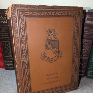 May include: A vintage book with a brown leather cover featuring a decorative border and a crest with a lion and shield. The book's title, "RALPH WALDO EMERSON," is printed in gold lettering. Other books are visible in the background.