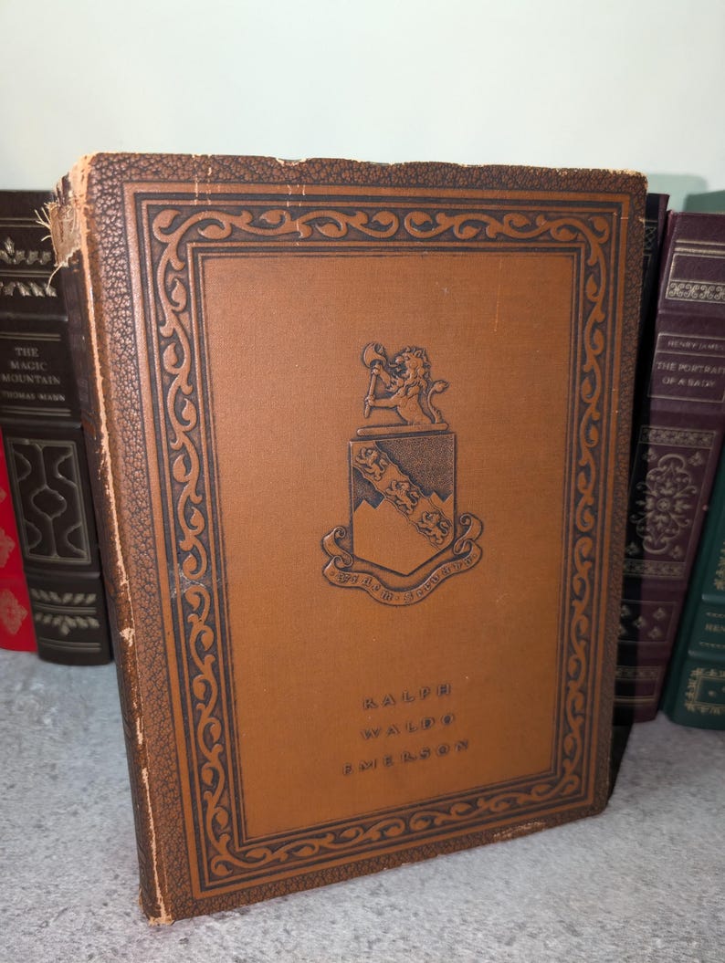 May include: A vintage book with a brown leather cover featuring a decorative border and a crest with a lion and shield. The book's title, "RALPH WALDO EMERSON," is printed in gold lettering. Other books are visible in the background.