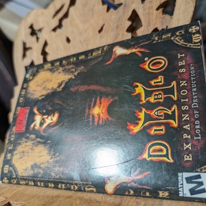 Op de afbeelding: Een Diablo II: Lord of Destruction uitbreidingsset videospeldoos met een zwarte en rode achtergrond. De doos toont een personage met een donkere kap en gloeiende rode ogen. De tekst "Diablo II" staat in een groot, gestileerd lettertype. De tekst "Expansion Set" staat in een kleiner lettertype onder de titel. De tekst "Lord of Destruction" staat in een kleiner lettertype onder de tekst van de uitbreidingsset. De doos heeft een "Mature"-classificatie van de ESRB.