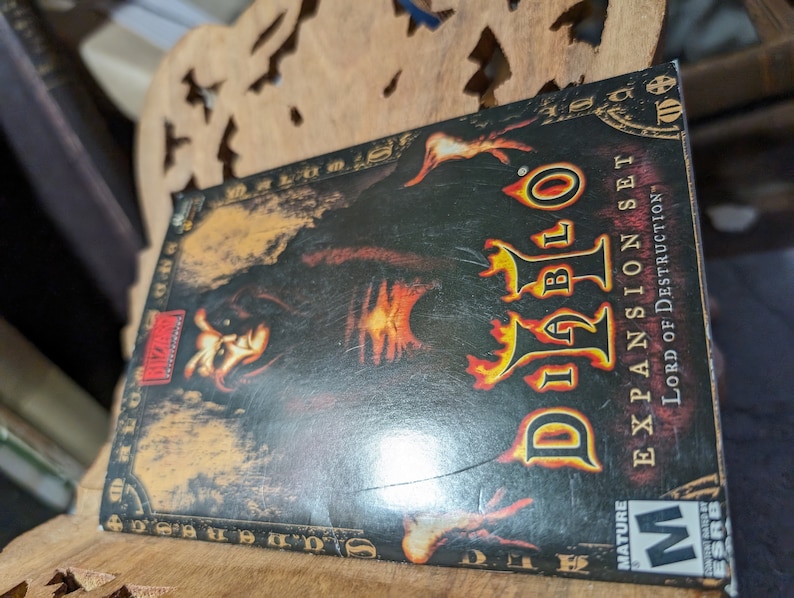 Op de afbeelding: Een Diablo II: Lord of Destruction uitbreidingsset videospeldoos met een zwarte en rode achtergrond. De doos toont een personage met een donkere kap en gloeiende rode ogen. De tekst "Diablo II" staat in een groot, gestileerd lettertype. De tekst "Expansion Set" staat in een kleiner lettertype onder de titel. De tekst "Lord of Destruction" staat in een kleiner lettertype onder de tekst van de uitbreidingsset. De doos heeft een "Mature"-classificatie van de ESRB.