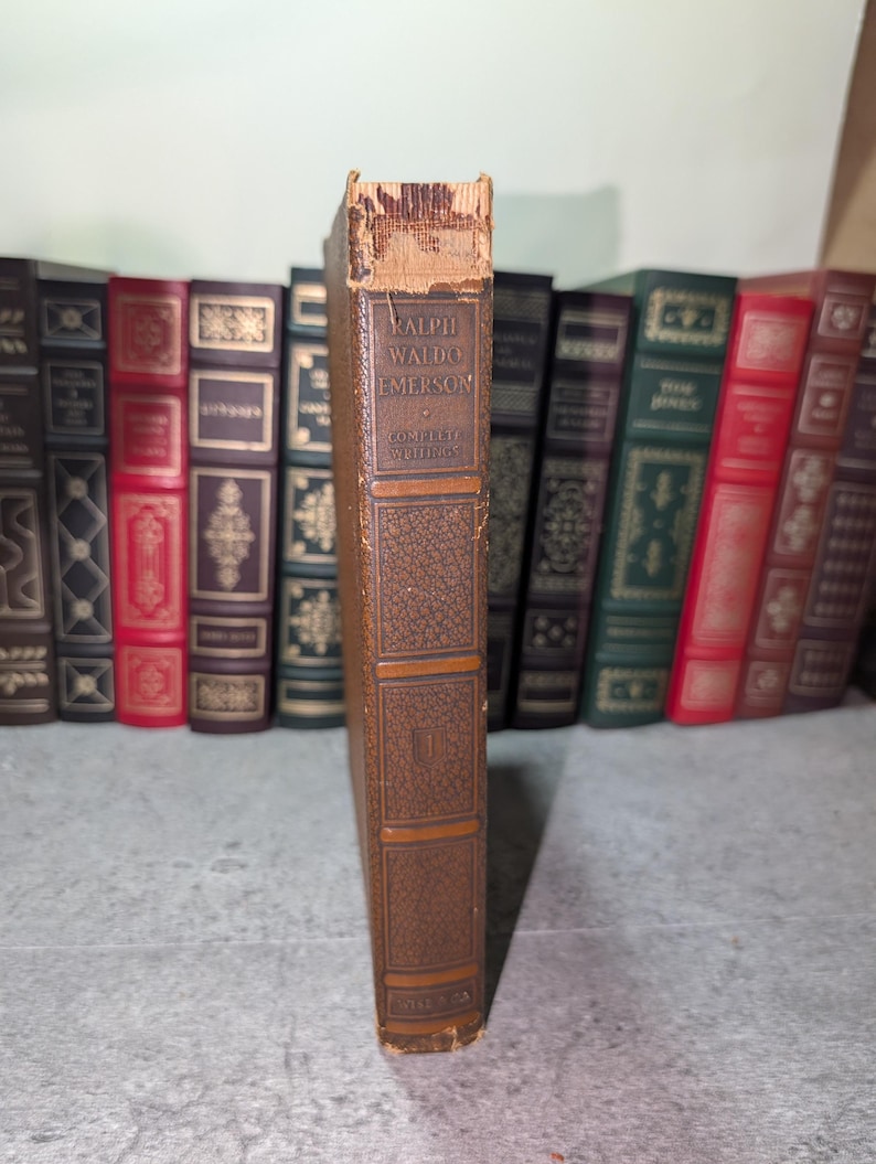 May include: A vintage brown book with the title "Ralph Waldo Emerson Complete Writings" visible on the spine. The book has a textured cover and is part of a collection of antique books.