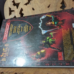 Op de afbeelding: De achterkant van een Diablo II videogamedoos. De doos is rood en zwart met een zilveren schild in het midden. Het schild heeft het Diablo II-logo erop. De achterkant van de doos vermeldt andere games van Blizzard Entertainment, waaronder Warcraft III, StarCraft en Brood War.