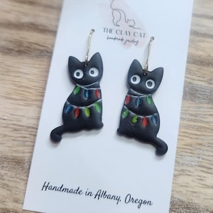 May include: A pair of black cat earrings with gold-colored hoops. Each cat is decorated with a string of colorful Christmas lights. The earrings are displayed on a white card with the text "THE CLAY CAT handmade jewelry".