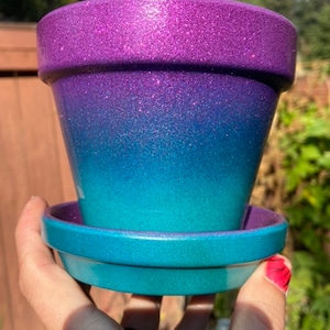 May include: A purple and blue ombre glitter planter with a matching saucer. The planter is made of terracotta and has a smooth, shiny finish.