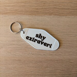 Può includere: Portachiavi bianco a forma di chiave di motel con il testo "shy extrovert" in lettere marroni.