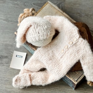 May include: A light pink knitted baby outfit set, including a hat with a long ear and a button-down onesie. The set is placed on a wooden box, with a small tag nearby. The outfit is made of soft, textured yarn.