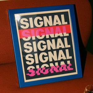 May include: A framed rectangular print featuring the word SIGNAL repeated five times in a bold, sans-serif font. The text is cream-colored against a dark background, with a horizontal red stripe and a pixelated pink glitch effect on the bottom.