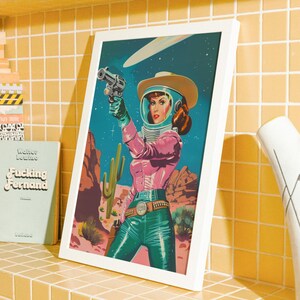 May include: A framed print featuring a retro-style illustration of a woman in a space helmet and cowboy attire, holding a gun. The artwork has a desert landscape background with a blue and pink color scheme.