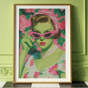 May include: A framed art print featuring a retro-style portrait of a woman with green skin, wearing pink cat-eye sunglasses and a pearl necklace. The background is a pink floral pattern, and the woman is wearing a floral dress.