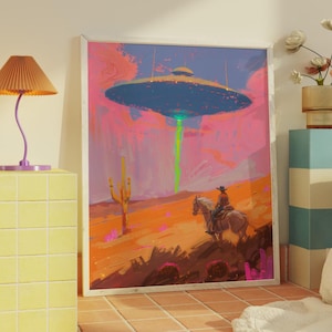 May include: Framed art print depicting a cowboy on horseback in a desert landscape, facing a hovering UFO. The artwork features a vibrant color palette with pink, orange, and blue hues. A green beam emanates from the spacecraft, adding a surreal element to the scene.
