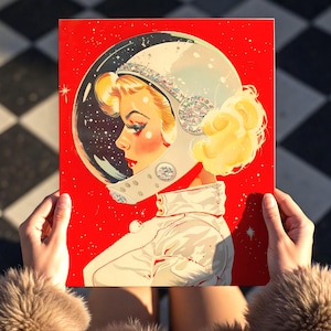 Op de afbeelding: Een retro-stijl print met een vrouw in een ruimtehelm tegen een felrode achtergrond. Het kunstwerk toont een blonde vrouw in profiel, gekleed in een wit ruimtepak en een helm versierd met juwelen. Het vizier van de helm onthult een sterrenstelsel.