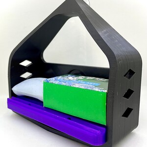 May include: A black plastic soap dish with a suction cup for hanging. The dish has a purple soap tray and is holding a green bar of soap and a white bar of soap.