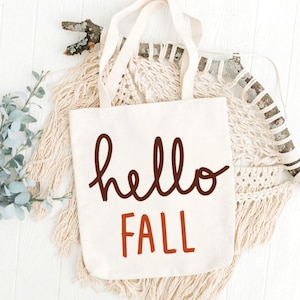 Peut inclure: Un sac en toile blanc avec le texte "hello fall" en lettres brunes et orange. Le sac est entouré d'une couverture en crochet blanc, d'une branche et d'une brindille d'eucalyptus.